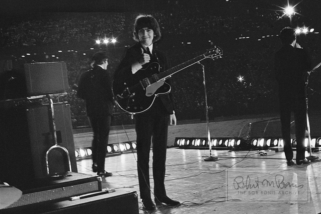 George Harrison, John Lennon and Paul McCartney, Metropolitan Stadium, Bloomington, MN, August 21, 1965 #4 George Harrison, wearing his beloved Gretsch Chet Atkins Country Gentleman model guitar, turns away from the audience toward the side of the stage and seeing  tour manager Bob Bonis standing there with his trusty Leica M3 camera, gives him a hearty thumbs-up.