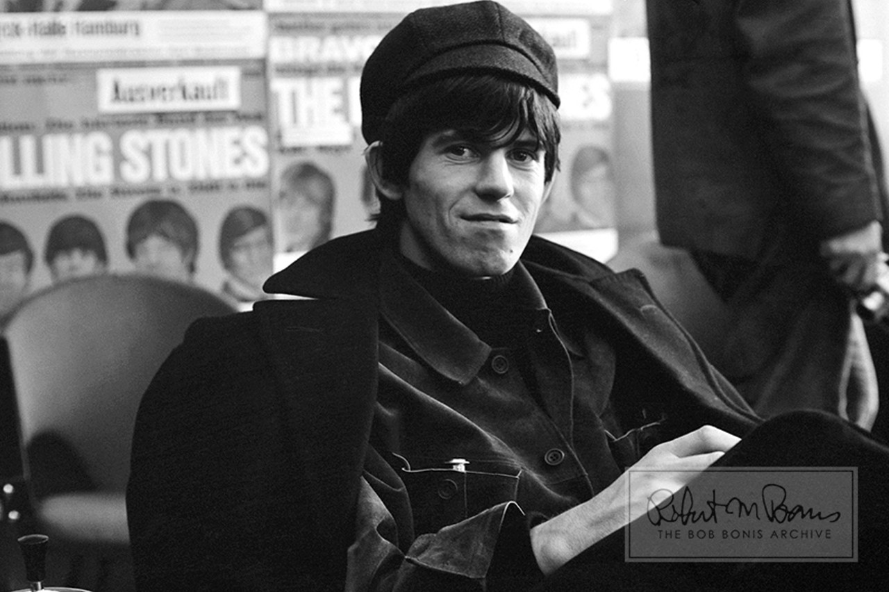 Keith Richards, Bild Zeitung Luncheon, Hamburg, West Germany, September 13, 1965 #1 1 Per BobBonis.com: "In a wonderfully meta moment, Keith Richards relaxes at a luncheon that was thrown by West German newspaper Bild Zeitung to make up for a mistake in an article that caused the Stones to threaten to cancel the rest of the tour.  The caption on a photo had misidentified Bill Wyman&#146;s girlfriend as Chrissie Shrimpton (the younger sister of model Jean Shrimpton), who was Mick Jagger&#146;s girlfriend, which caused manager Andrew Loog Oldham to force the paper to print a retraction.  Bob Bonis accompanied the Stones on their brief, five-city tour of West Germany &#150; and the tour was a riot-filled affair. Former Stones bassist Bill Wyman recalls 'police dogs everywhere' and crowds 'estimated at between 21,000 and 23,000.' In this photo, behind Keith is a wall plastered with German Rolling Stones posters, sponsored by Bild Zeitung&#146;s publication BRAVO. After rioting broke out at the West Berlin show on September 15, the East and West German press reprinted descriptions from Bild Zeitung of girls throwing off their underwear in ecstasy &#150; in an effort to censor American and British influences, just as they had done with Elvis Presley in 1956."