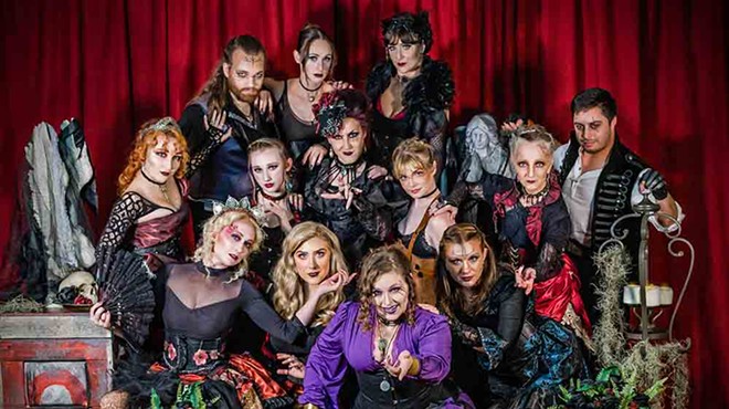 A huge group of spooky steampunk performers, storytellers, dancers, jugglers, puppeteers, etc. are posed in dark, ornate clothing in front of a red velvet curtain.