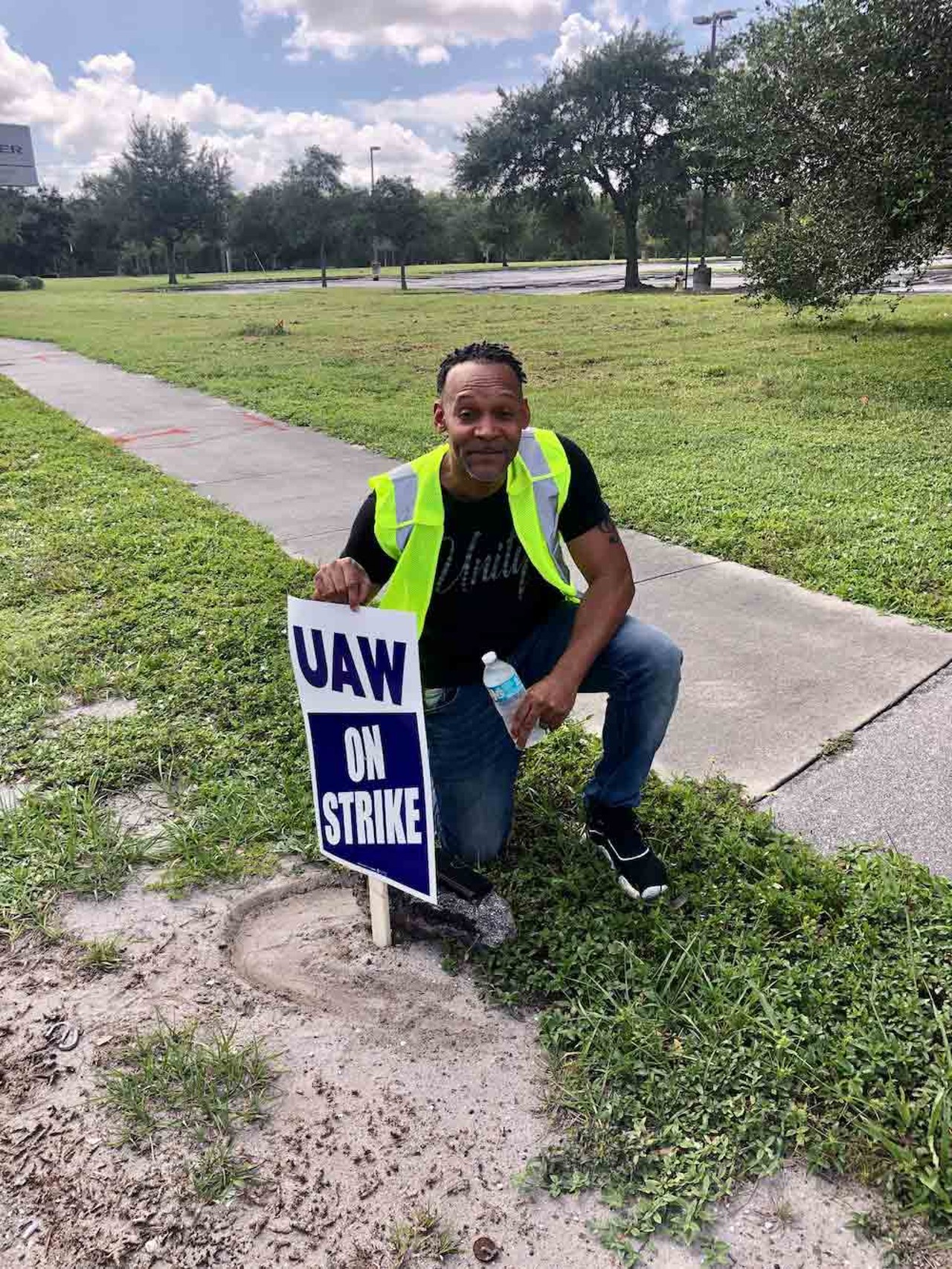 Keith Phelps, a UAW member and auto worker, kneels next to a UAW picket sign outside of a Chrysler/Stellantis parts depot in Orlando on Sept. 22, 2023.
