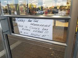 A handwritten sign on the front door of Belk explaining the store is closed "due to Mother Nature." Workers are seen just inside with giant dryers working to clean up a recent leak.